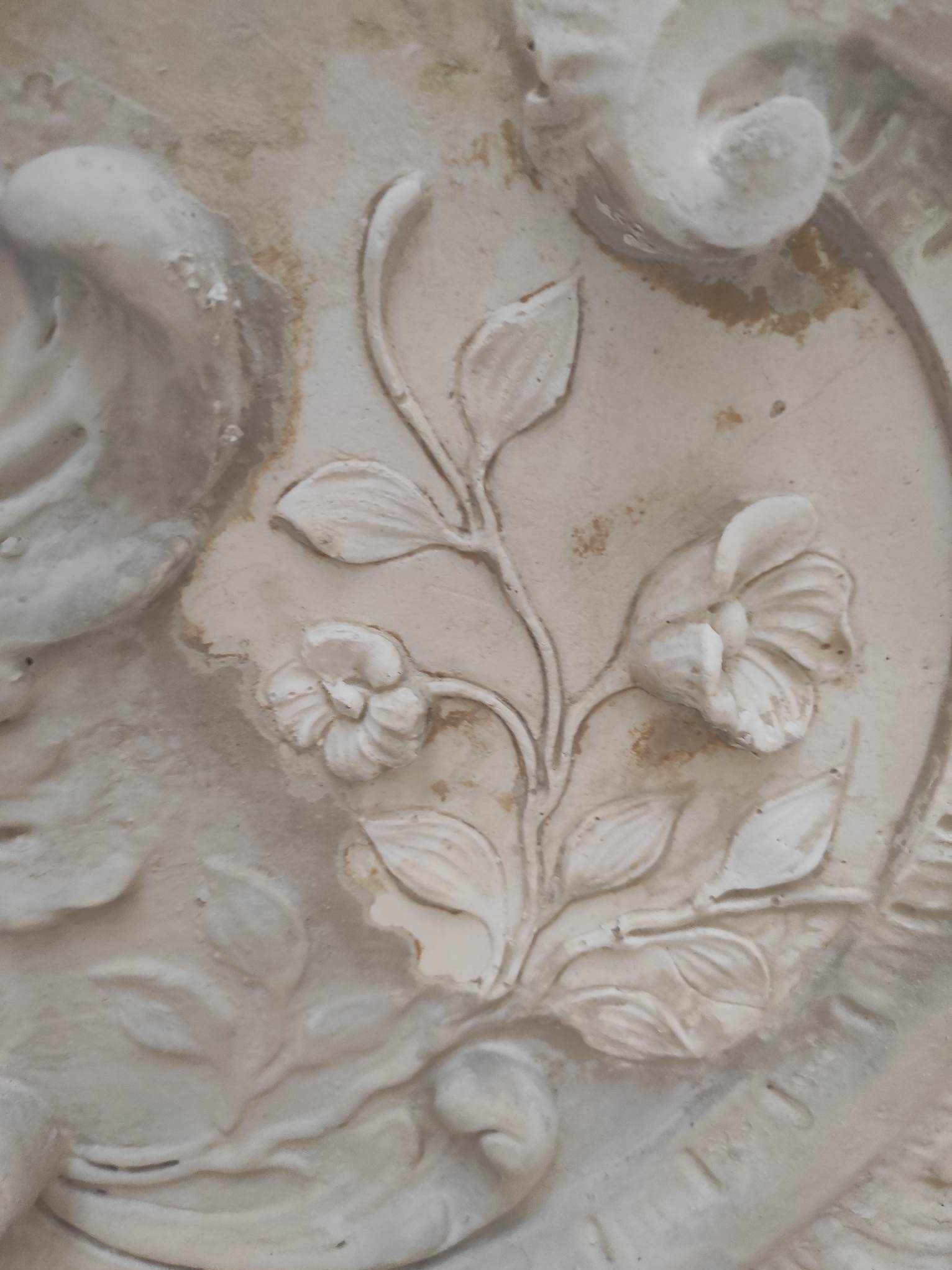 Detal płaskorzeźby z motywem kwiatowym na jasnej elewacji, widoczne ślady patyny i delikatne uszkodzenia, podkreślające wiekowy charakter dekoracji.