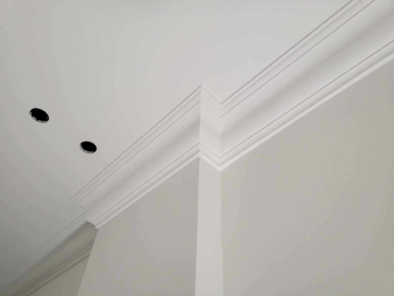 Białe, eleganckie listwy dekoracyjne na styku ściany i sufitu, z widocznymi otworami na oświetlenie punktowe, tworzące estetyczne wykończenie wnętrza.