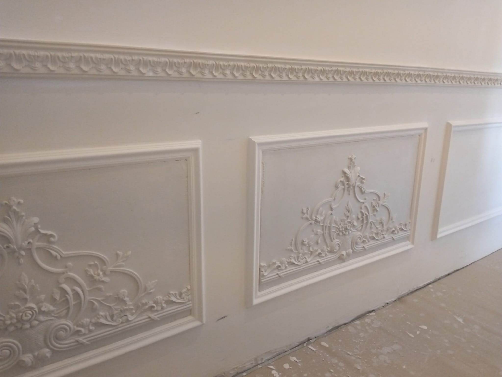 Biała ściana z dekoracyjnymi, gipsowymi panelami i listwami w stylu klasycznym. Detal wykończenia wnętrza po renowacji, widoczne ślady prac na podłodze.