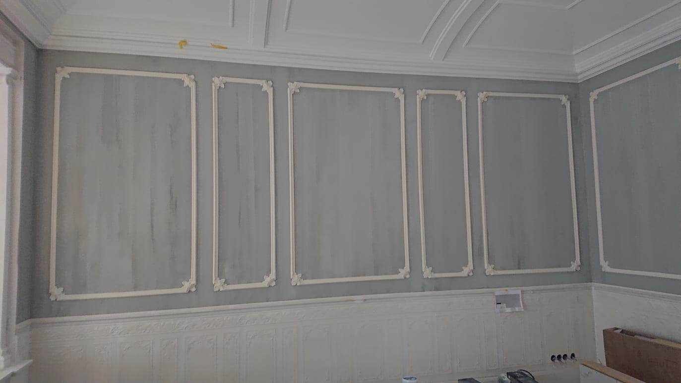 Szara ściana z dekoracyjnymi, białymi panelami i listwami. Klasyczny wystrój wnętrza z subtelnymi zdobieniami.