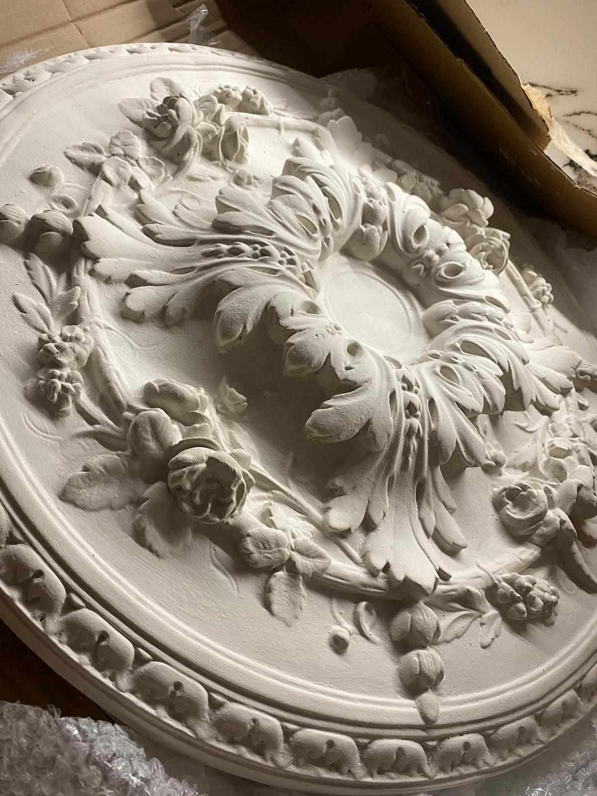 Okrągły, gipsowy element dekoracyjny z motywem roślinnym, prawdopodobnie rozeta sufitowa, w odcieniach bieli, leżący na folii bąbelkowej i kartonie.