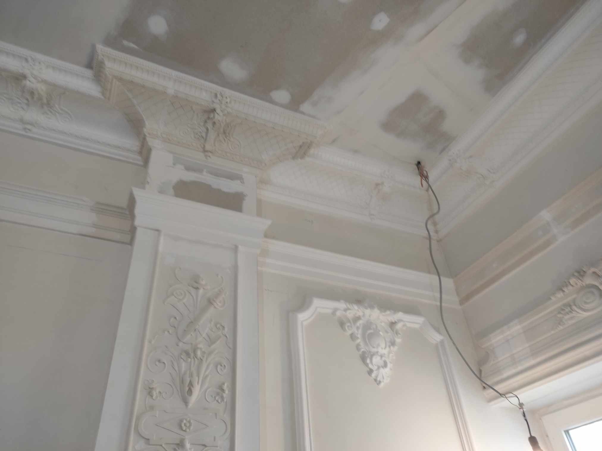 Białe, zdobione sztukaterie na ścianie i suficie w trakcie prac wykończeniowych. Widoczne ślady szpachlowania na suficie. Dekoracyjne detale architektoniczne.
