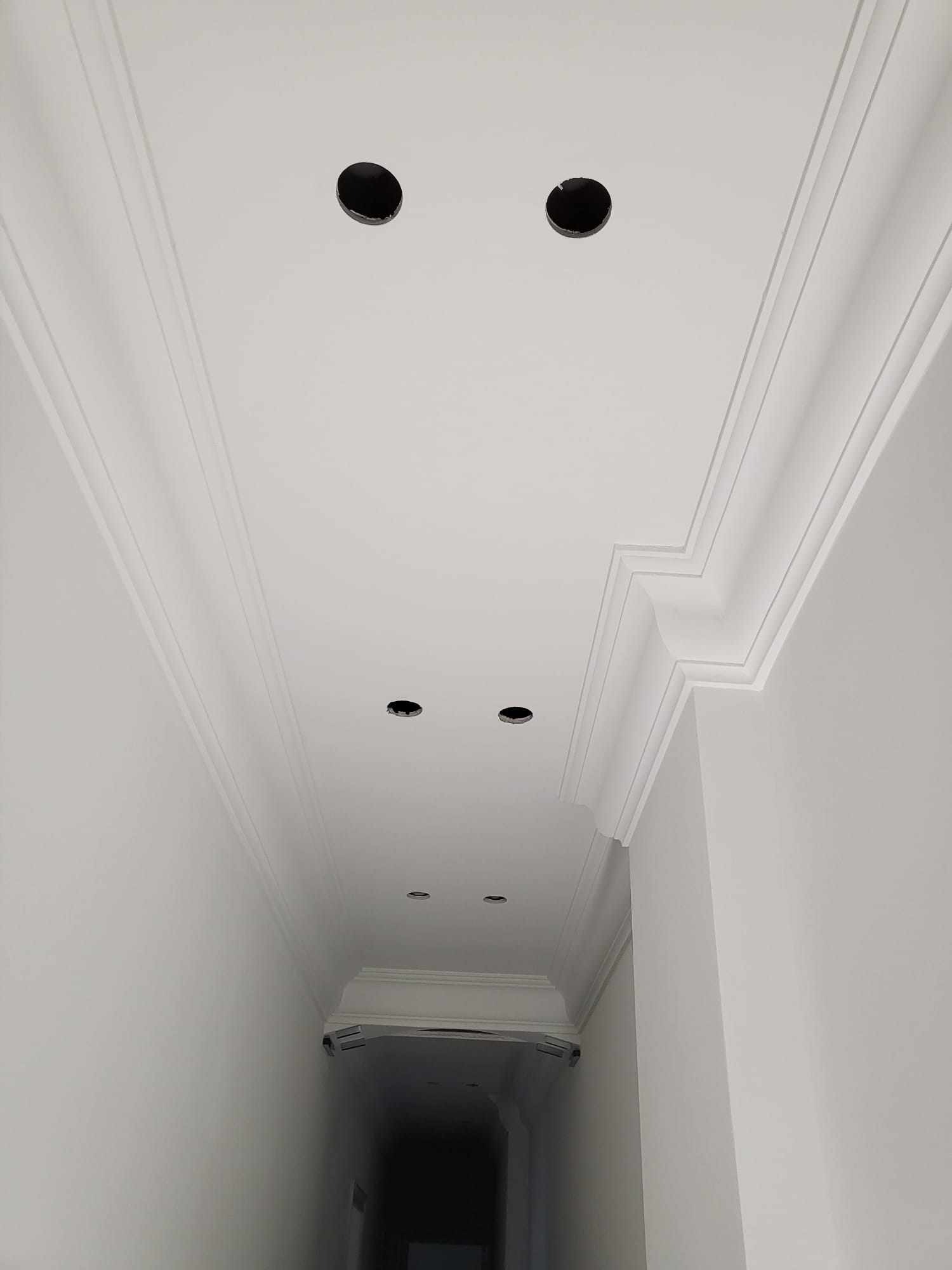 Biały sufit z ozdobnymi listwami i otworami na oświetlenie, widok z dołu w korytarzu. Minimalistyczny design, świeżo po remoncie.