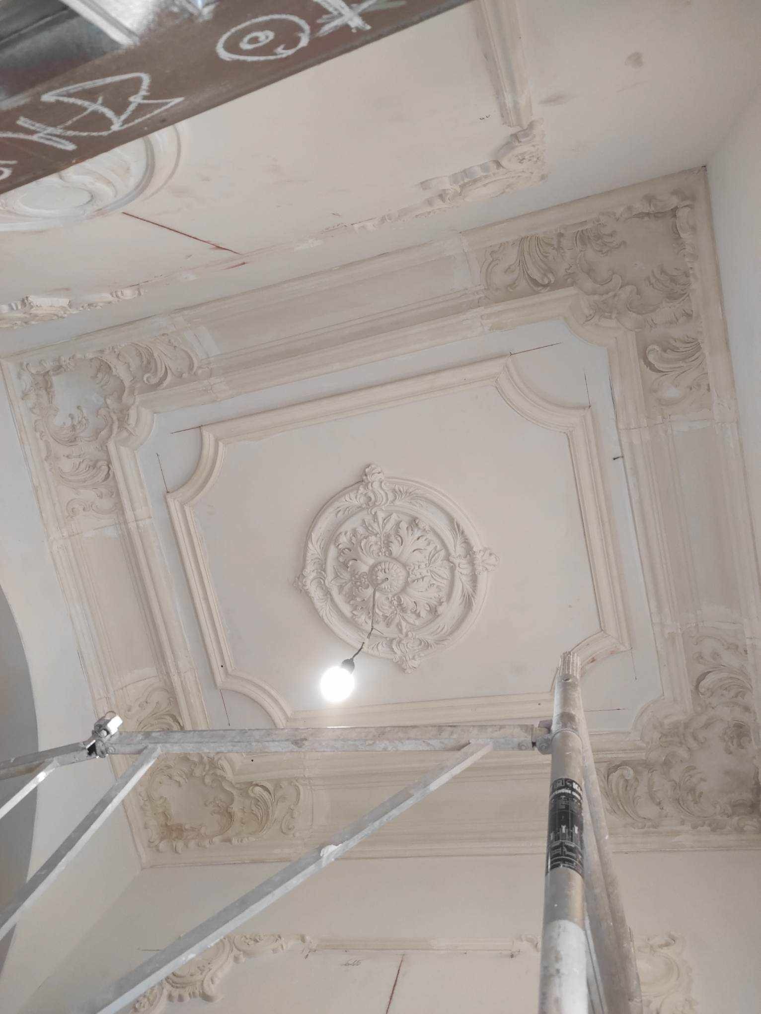 Biały sufit z dekoracyjnymi sztukateriami w trakcie renowacji, widoczny fragment rusztowania. Centralnie umieszczona rozeta z ornamentami. Oświetlenie punktowe.