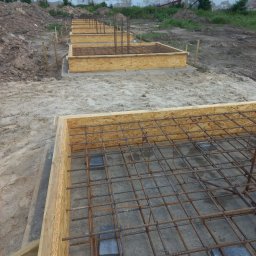 Dremex - Przygotowane szalunki zbrojeniowe pod fundamenty na piaszczystym podłożu. Widoczne drewniane ramy i siatka zbrojeniowa. W tle budynek gospodarczy i hałda ziemi.