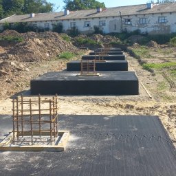 Dremex - Przygotowane, zaizolowane fundamenty pod budynek z wystającymi zbrojeniami, na piaszczystym terenie. W tle budynek gospodarczy i drzewa.