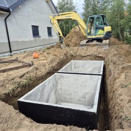 Dremex - Dwa betonowe zbiorniki w wykopie, żółta koparka Yanmar w tle, obok budynek w trakcie budowy. Widok z góry na dół.