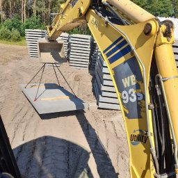 Dremex - Żółta koparka Komatsu podnosi betonową płytę za pomocą łańcuchów na placu budowy. W tle stosy płyt i drzewa.
