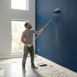 Marta Witczak - Malarz z wałkiem teleskopowym maluje na granatowo ścianę w jasnym wnętrzu. Podłoga zabezpieczona folią malarską, widoczna drabina i okno.