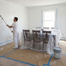 Marta Witczak - Malowanie ścian w pokoju: malarz z wałkiem, zabezpieczone meble i podłoga. Widoczne taśmy malarskie. Świeżo pomalowana ściana w jasnym odcieniu bieli.