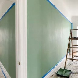 Marta Witczak - Korytarz w trakcie malowania na pistacjowy kolor, zabezpieczony taśmą malarską i folią. Widoczna drabina i kuweta z wałkiem. Świeżo pomalowana ściana.
