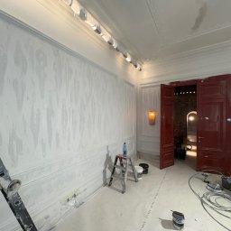 TOP HOUSE EŁK SPÓŁKA Z OGRANICZONĄ ODPOWIEDZIALNOŚCIĄ - Wnętrze w trakcie prac wykończeniowych: ściany z dekoracyjną fakturą, listwy ozdobne, drabina, narzędzia malarskie. Widoczne czerwone drzwi i oświetlenie.