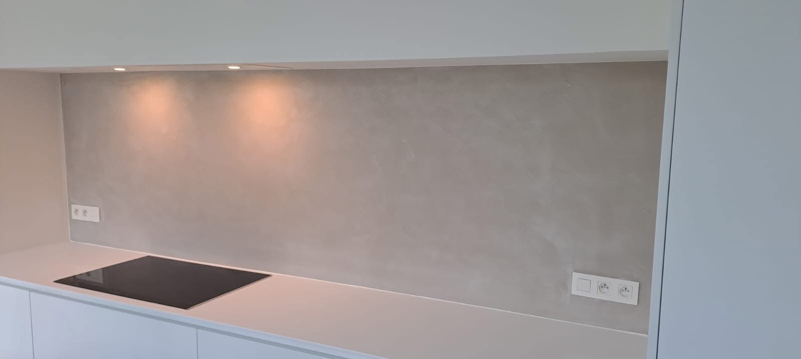 Minimalistyczna kuchnia z szarą ścianą o fakturze betonu, oświetlona punktowo. Widoczne gniazdka elektryczne i płyta indukcyjna. Nowoczesny design.