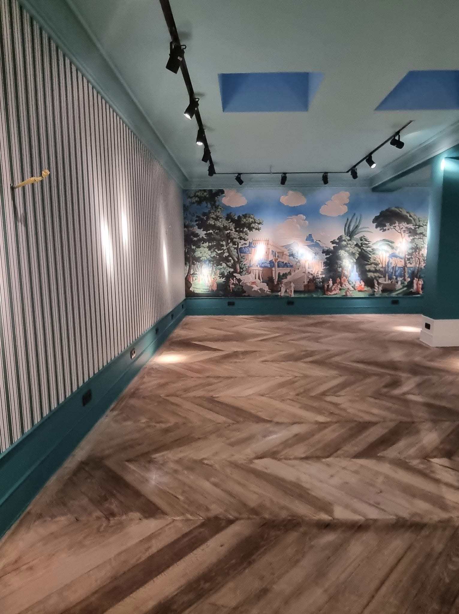 Wnętrze po renowacji: parkiet w jodełkę, tapeta w pionowe pasy i malowidło na ścianie. Oświetlenie punktowe podkreśla detale. Wykończenie w odcieniach zieleni i bieli.