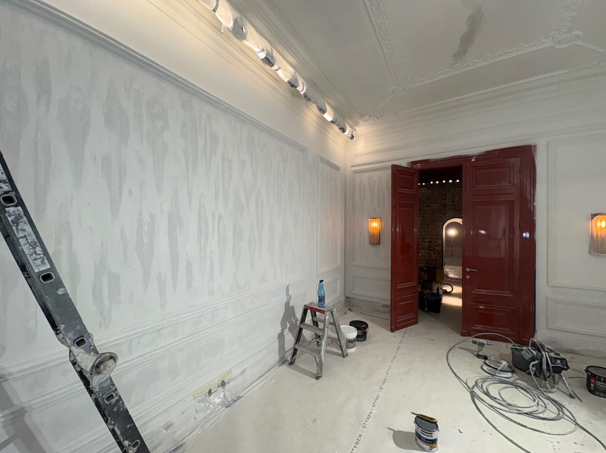 Wnętrze w trakcie prac wykończeniowych: ściany z dekoracyjną fakturą, listwy ozdobne, drabina, narzędzia malarskie. Widoczne czerwone drzwi i oświetlenie.