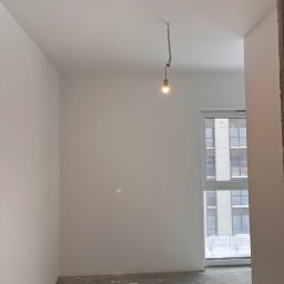 MG USŁUGI REMONTOWO BUDOWLANE MATEUSZ GRZYB - Wnętrze pokoju po malowaniu ścian na biało, widoczne okno z widokiem na budynek. Na suficie prowizoryczna żarówka. Podłoga surowa, przygotowana do dalszych prac.