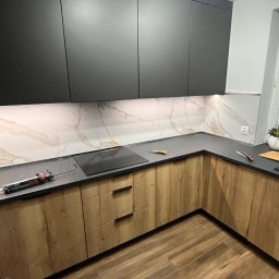 MG USŁUGI REMONTOWO BUDOWLANE MATEUSZ GRZYB - Nowoczesna kuchnia z drewnianymi szafkami dolnymi i czarnymi górnymi, marmurowy backsplash, czarny blat, drewniana podłoga. Widoczne narzędzia na blacie.