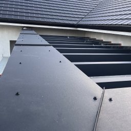 MG USŁUGI REMONTOWO BUDOWLANE MATEUSZ GRZYB - Czarne, metalowe obróbki blacharskie na dachu domu, mocowane na nity. Widok z dołu, fragment dachu z czarną dachówką w tle.