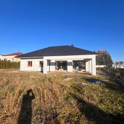 MG USŁUGI REMONTOWO BUDOWLANE MATEUSZ GRZYB - Nowoczesny, parterowy dom jednorodzinny z ciemnym dachem i dużymi oknami, w stanie surowym zamkniętym, na tle błękitnego nieba i zarośniętej działki. Widoczny cień fotografa.