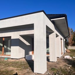 MG USŁUGI REMONTOWO BUDOWLANE MATEUSZ GRZYB - Nowoczesny dom jednorodzinny z jasną elewacją i ciemnym dachem w słoneczny dzień. Widoczne kolumny podtrzymujące zadaszenie i okna z drewnianą stolarką. Teren wokół domu w trakcie prac.