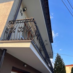 Elegancki balkon z kutą balustradą, świeżo po renowacji. Widoczne detale wykończenia elewacji i oświetlenie zewnętrzne. Ujęcie z dołu, podkreślające bryłę budynku.
