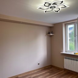 MG USŁUGI REMONTOWO BUDOWLANE MATEUSZ GRZYB - Jasne wnętrze pokoju po remoncie: drewnopodobne panele, beżowe ściany z listwą, nowoczesna lampa sufitowa w kształcie kwiatu, okno z grzejnikiem.