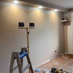 MG USŁUGI REMONTOWO BUDOWLANE MATEUSZ GRZYB - Świeżo pomalowane wnętrze z drabiną i lampą budowlaną. Widoczne detale wykończenia sufitu i gładka powierzchnia ściany w odcieniach beżu. Przygotowanie do dalszych prac.