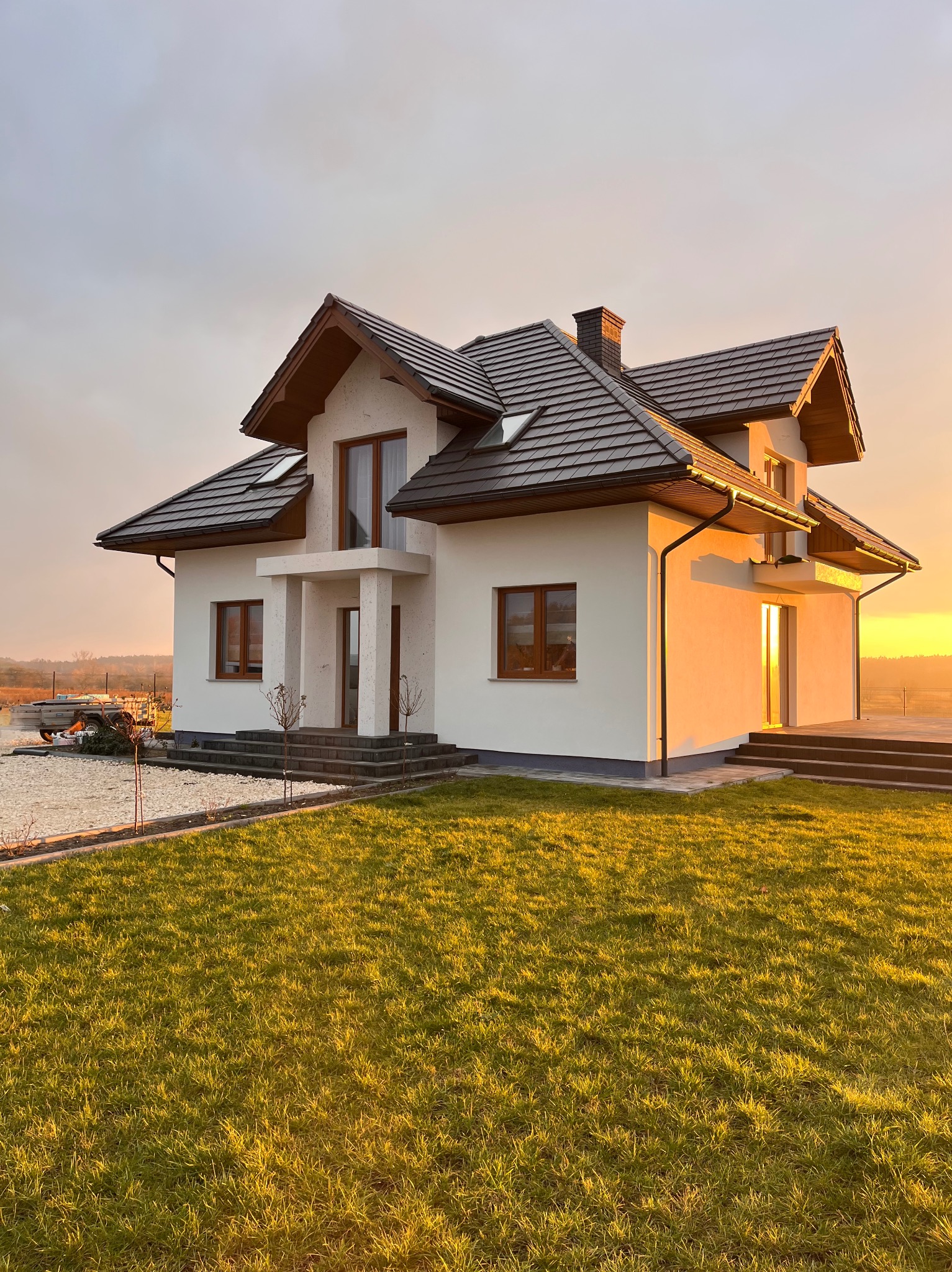 Elewacja domu jednorodzinnego w ciepłym świetle zachodzącego słońca. Widoczne brązowe okna i dachówka, biała elewacja oraz zadbany trawnik.