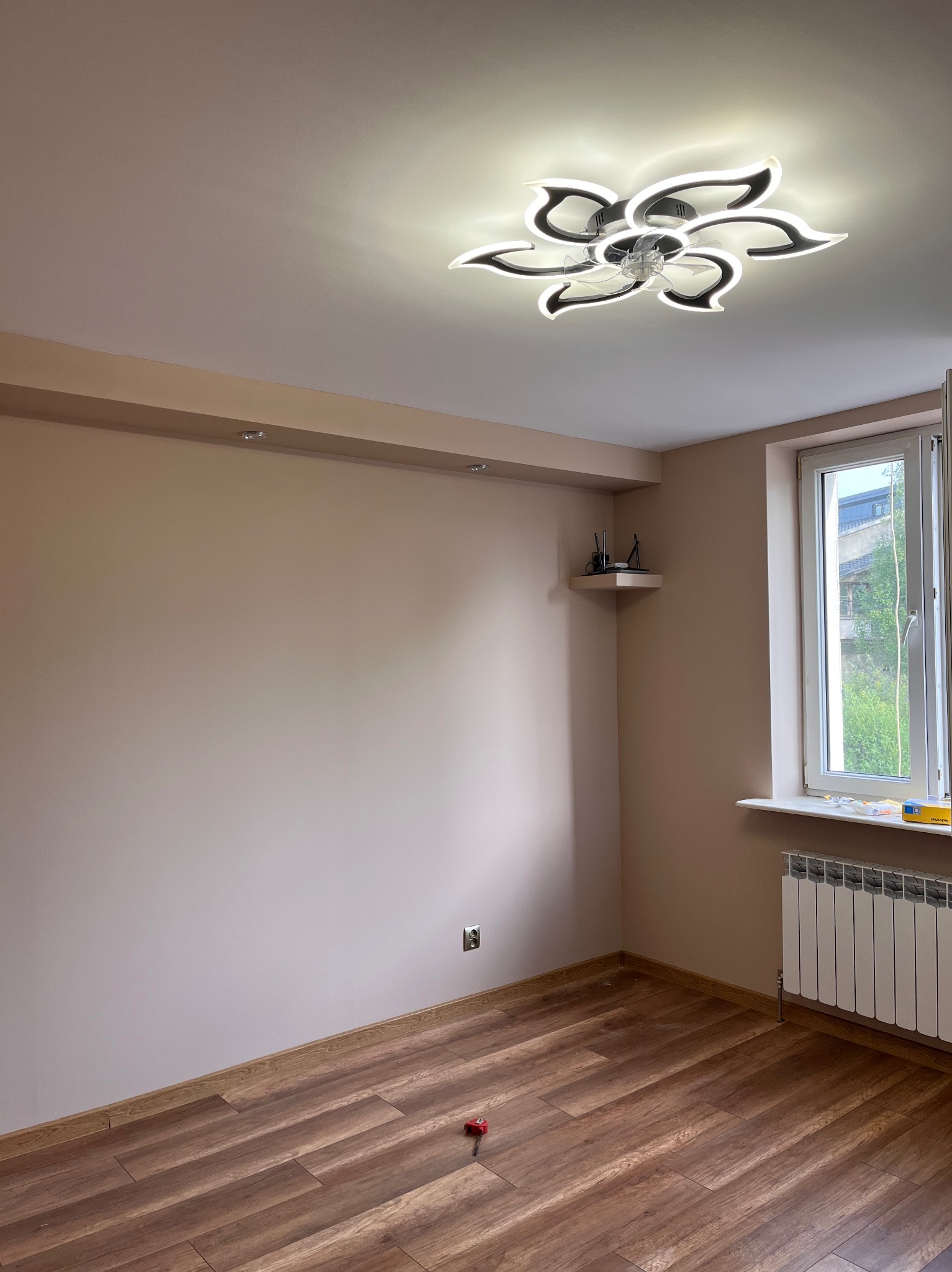 Jasne wnętrze pokoju po remoncie: drewniana podłoga, beżowe ściany, nowoczesna lampa sufitowa w kształcie kwiatu, okno z grzejnikiem. Widoczny fragment półki.