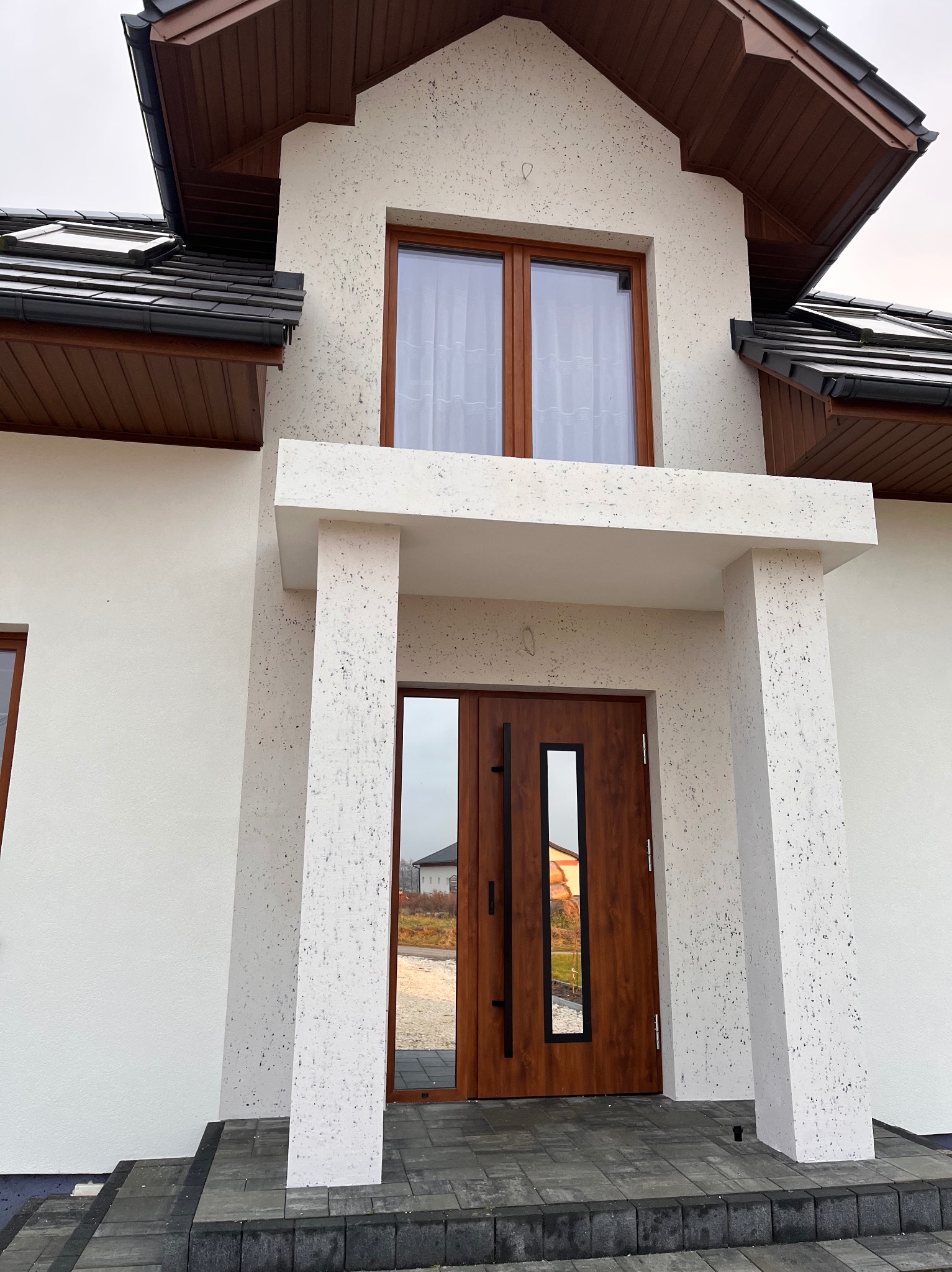 Elewacja domu jednorodzinnego z jasnym tynkiem barankiem, drewnianą stolarką okienną i drzwiami z przeszkleniem. Widoczne schody wejściowe z szarej kostki.