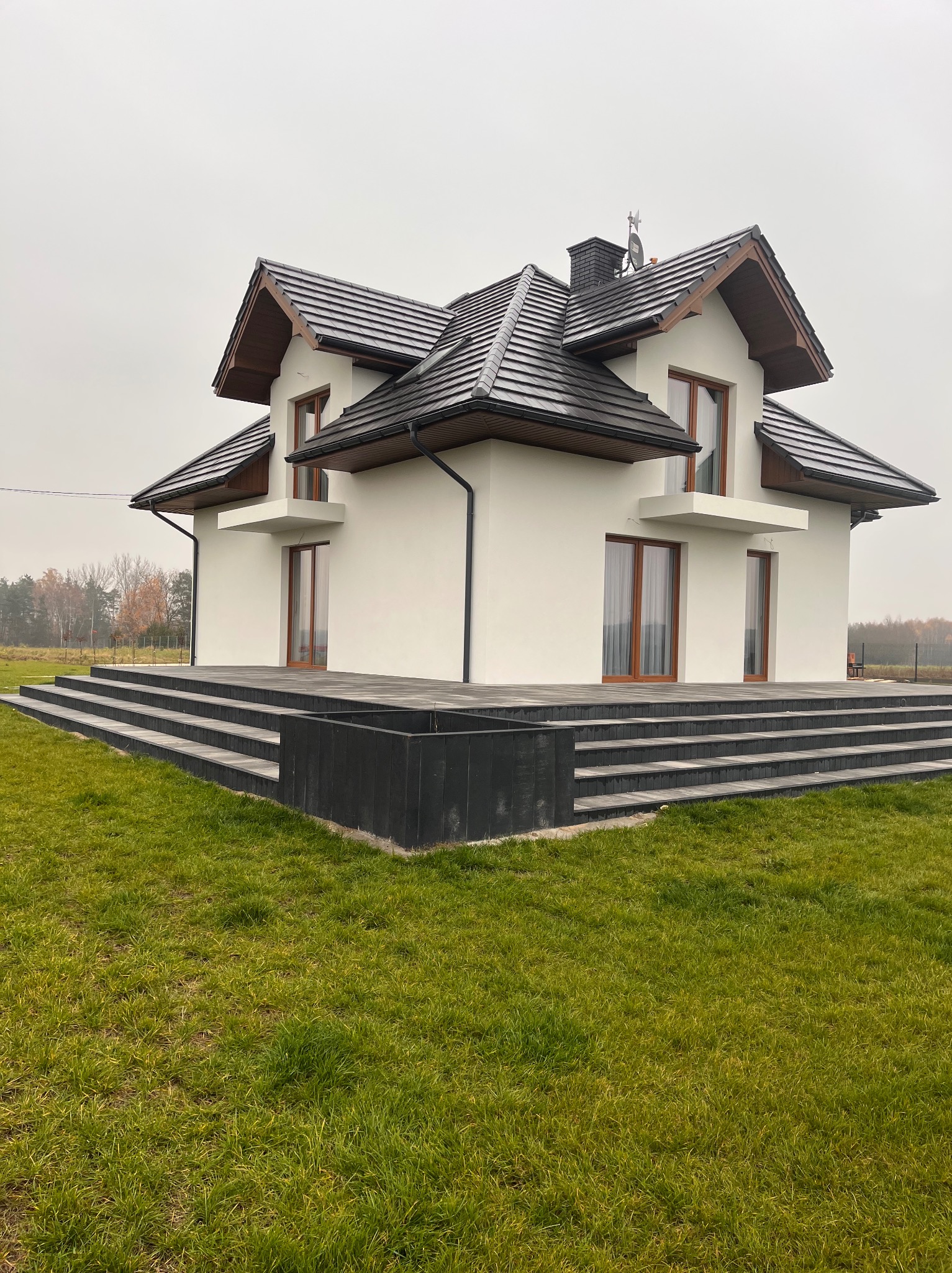 Nowoczesny dom jednorodzinny z białymi ścianami, ciemną dachówką i drewnianymi oknami, otoczony zielonym trawnikiem w pochmurny dzień. Widoczne schody wejściowe.
