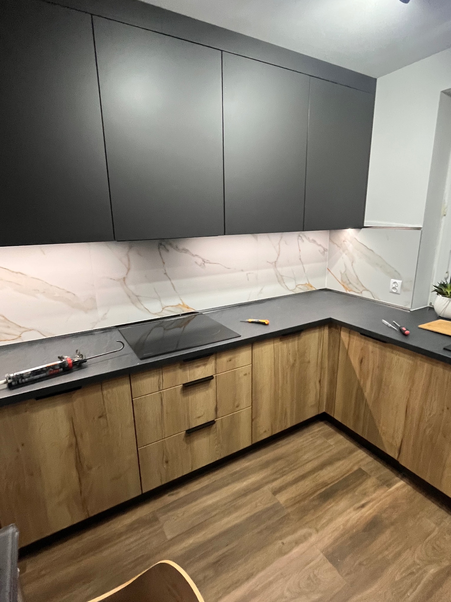 Nowoczesna kuchnia z drewnianymi szafkami dolnymi i czarnymi górnymi, marmurowy backsplash, czarny blat, drewniana podłoga. Widoczne narzędzia na blacie.