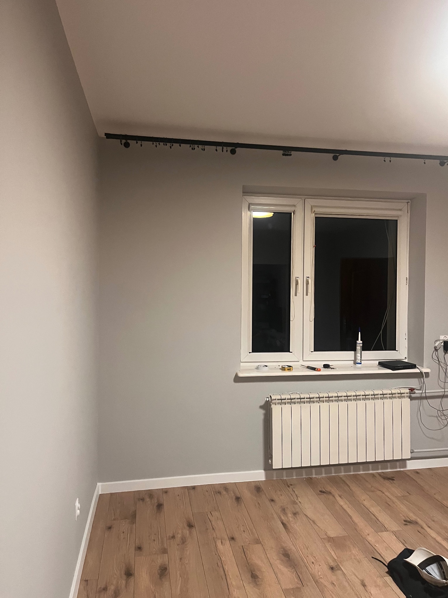Szare wnętrze po remoncie: drewniana podłoga, białe okno z grzejnikiem, listwa oświetleniowa na suficie. Widoczne narzędzia na parapecie.