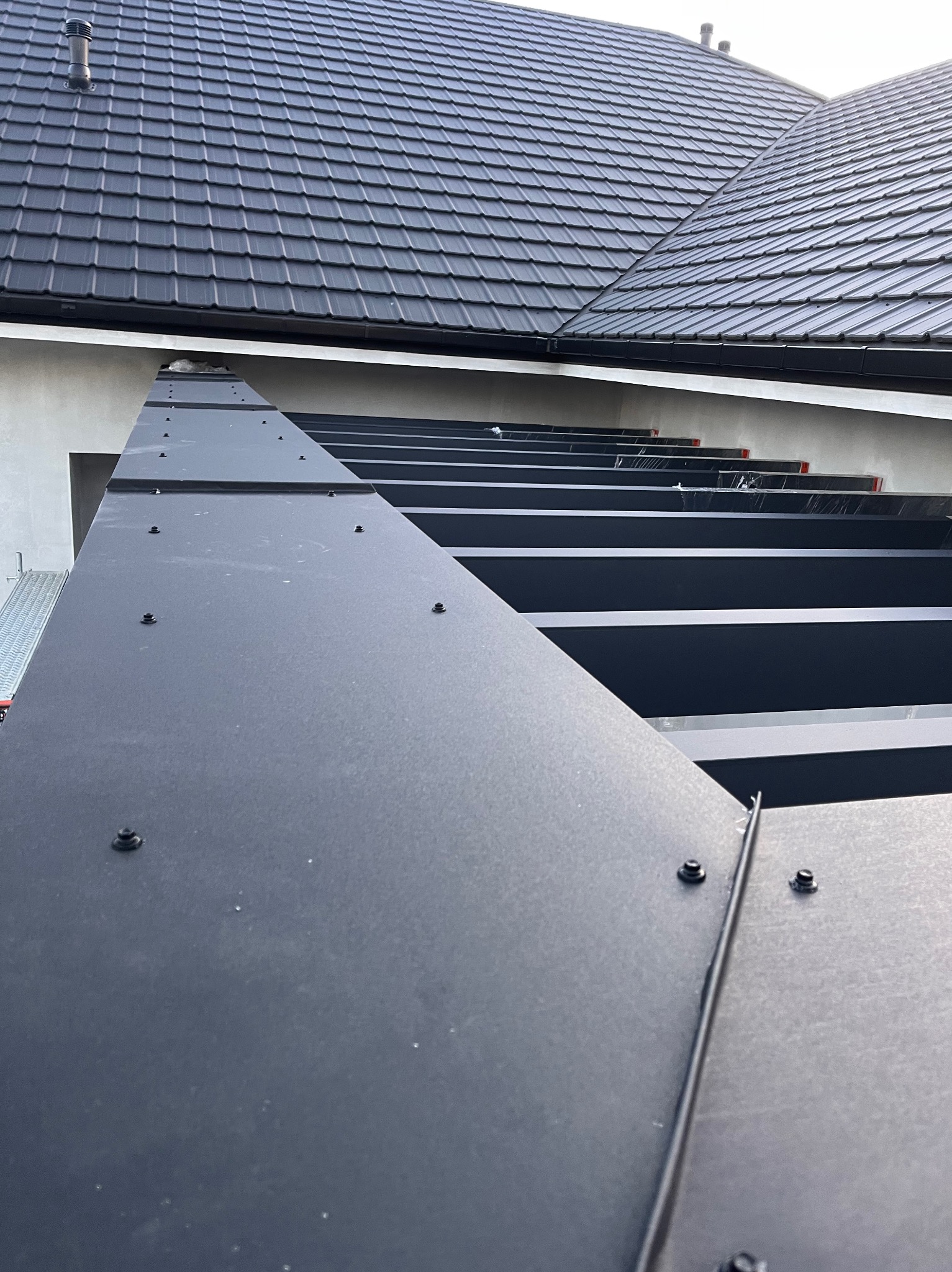 Czarne, metalowe obróbki blacharskie na dachu domu, mocowane na nity. Widok z dołu, fragment dachu z czarną dachówką w tle.