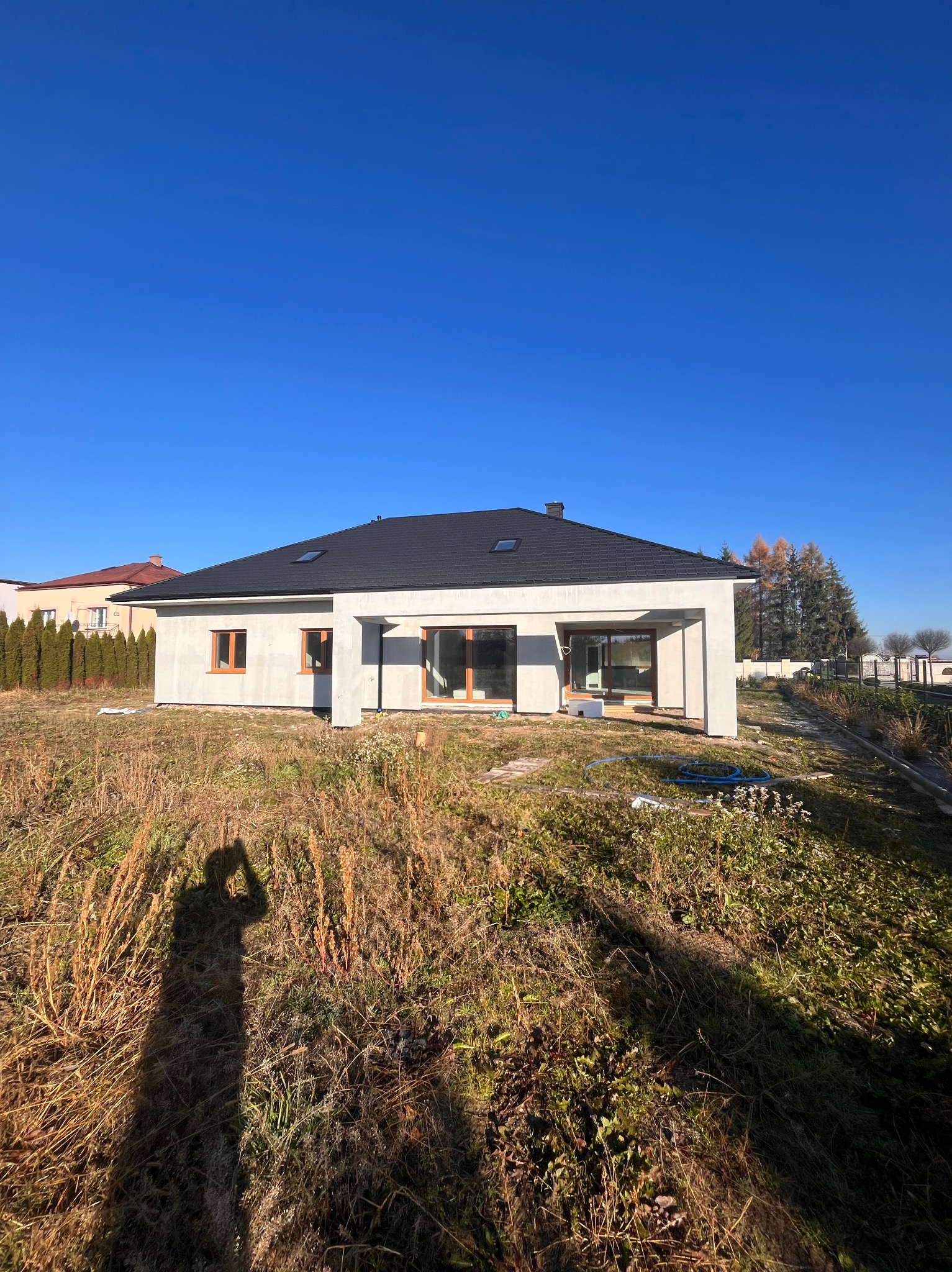 Nowoczesny, parterowy dom jednorodzinny z ciemnym dachem i dużymi oknami, w stanie surowym zamkniętym, na tle błękitnego nieba i zarośniętej działki. Widoczny cień fotografa.