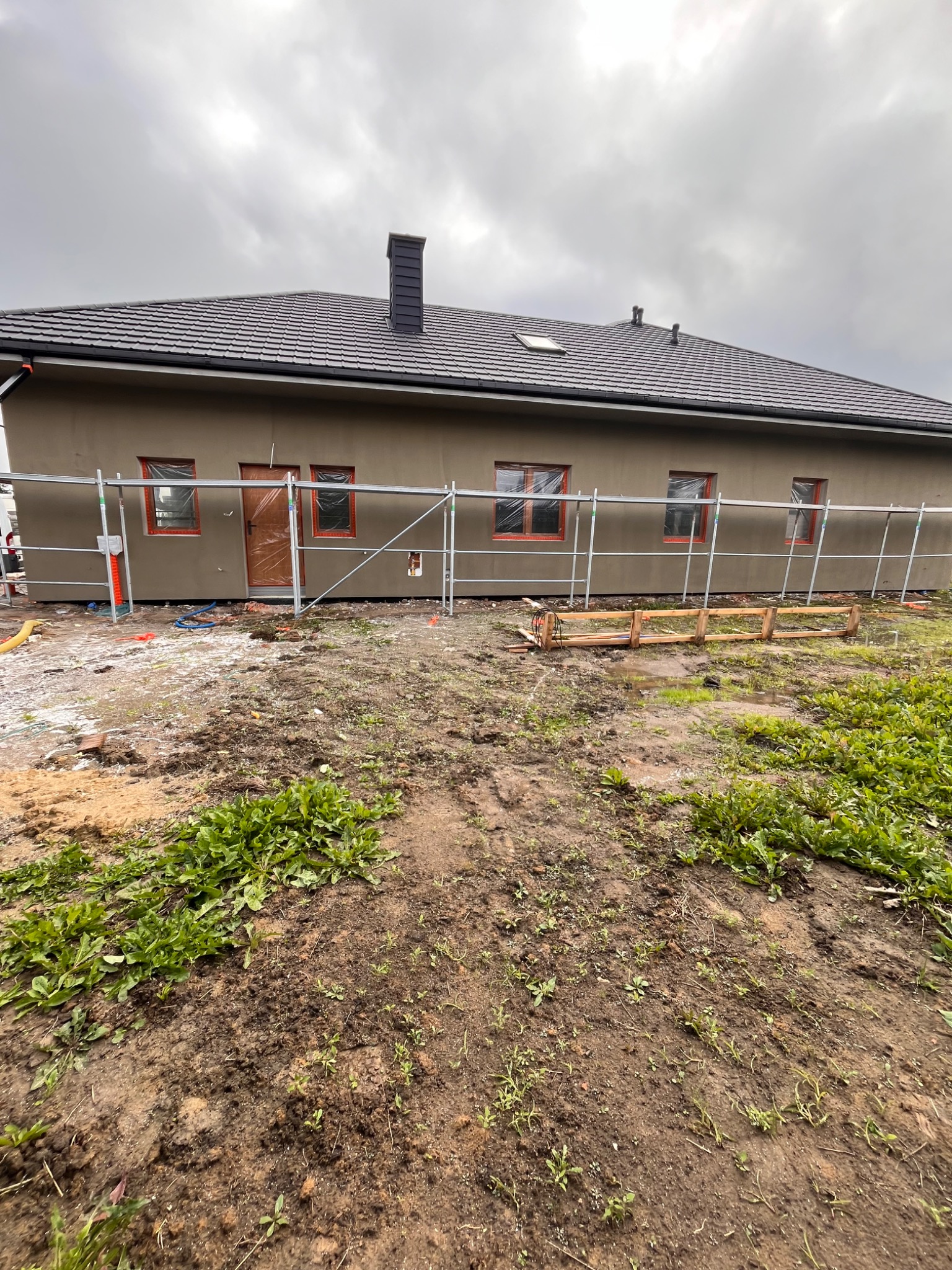 Dom jednorodzinny w trakcie prac elewacyjnych, z widocznym rusztowaniem i zabezpieczonymi oknami. Tynk w kolorze beżowym, dach z ciemnej dachówki.