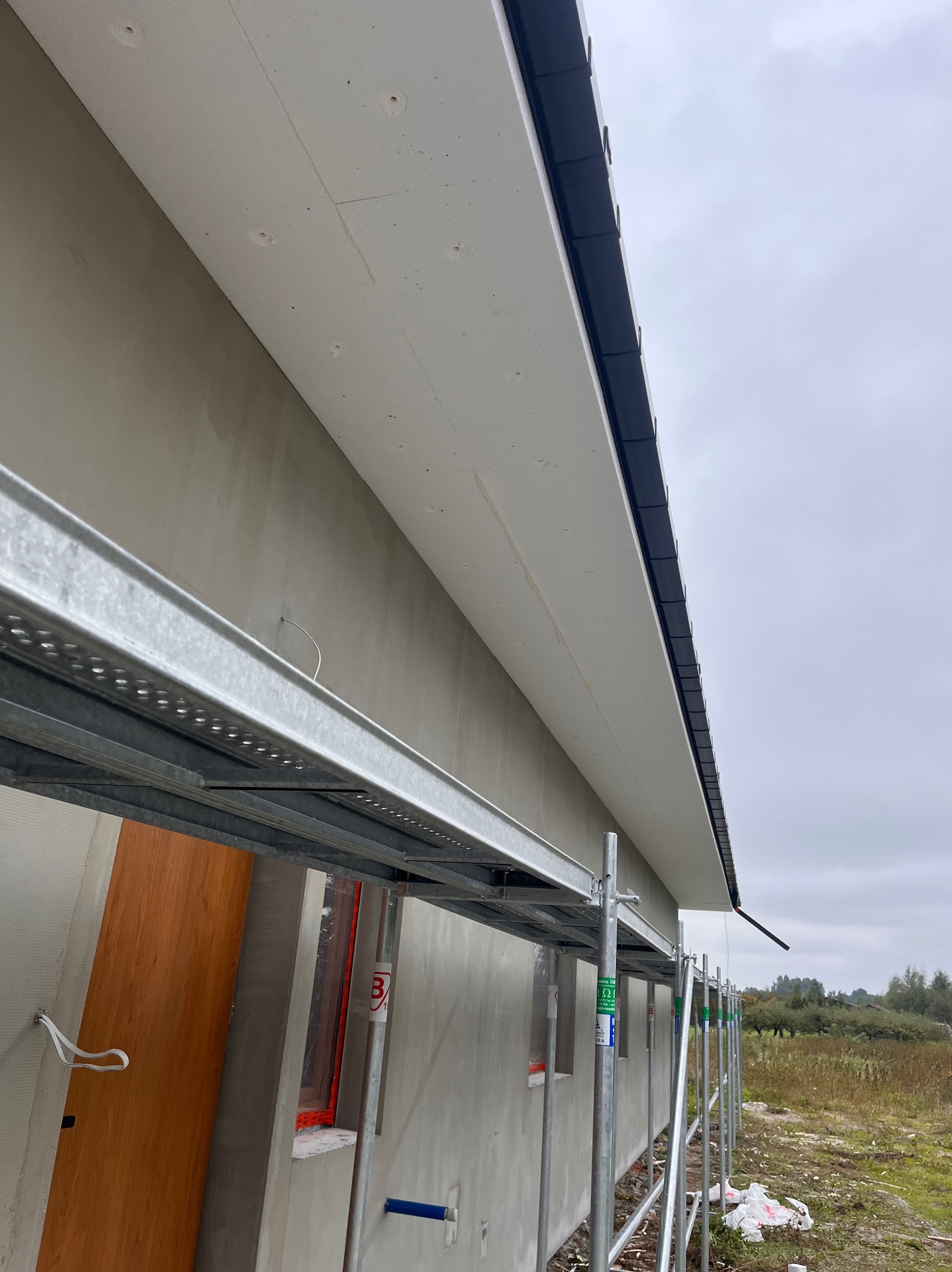 Elewacja budynku w trakcie prac wykończeniowych, widoczne rusztowania, ocieplenie i podbitka dachowa. Ujęcie z dołu, pochmurne niebo w tle.