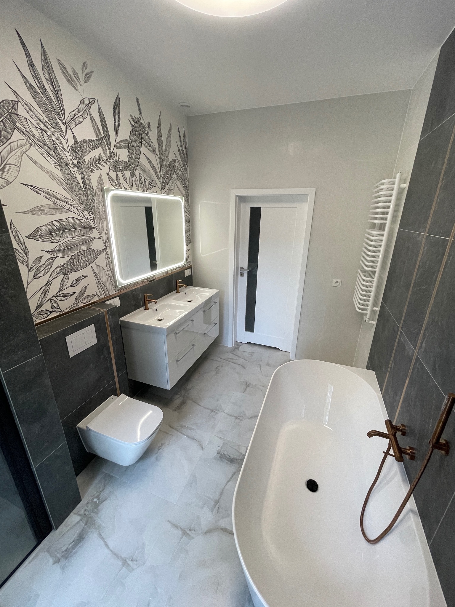 Nowoczesna łazienka z wanną, umywalką z podświetlanym lustrem i toaletą. Na ścianie tapeta z motywem roślinnym. Uwagę przykuwają miedziane baterie.