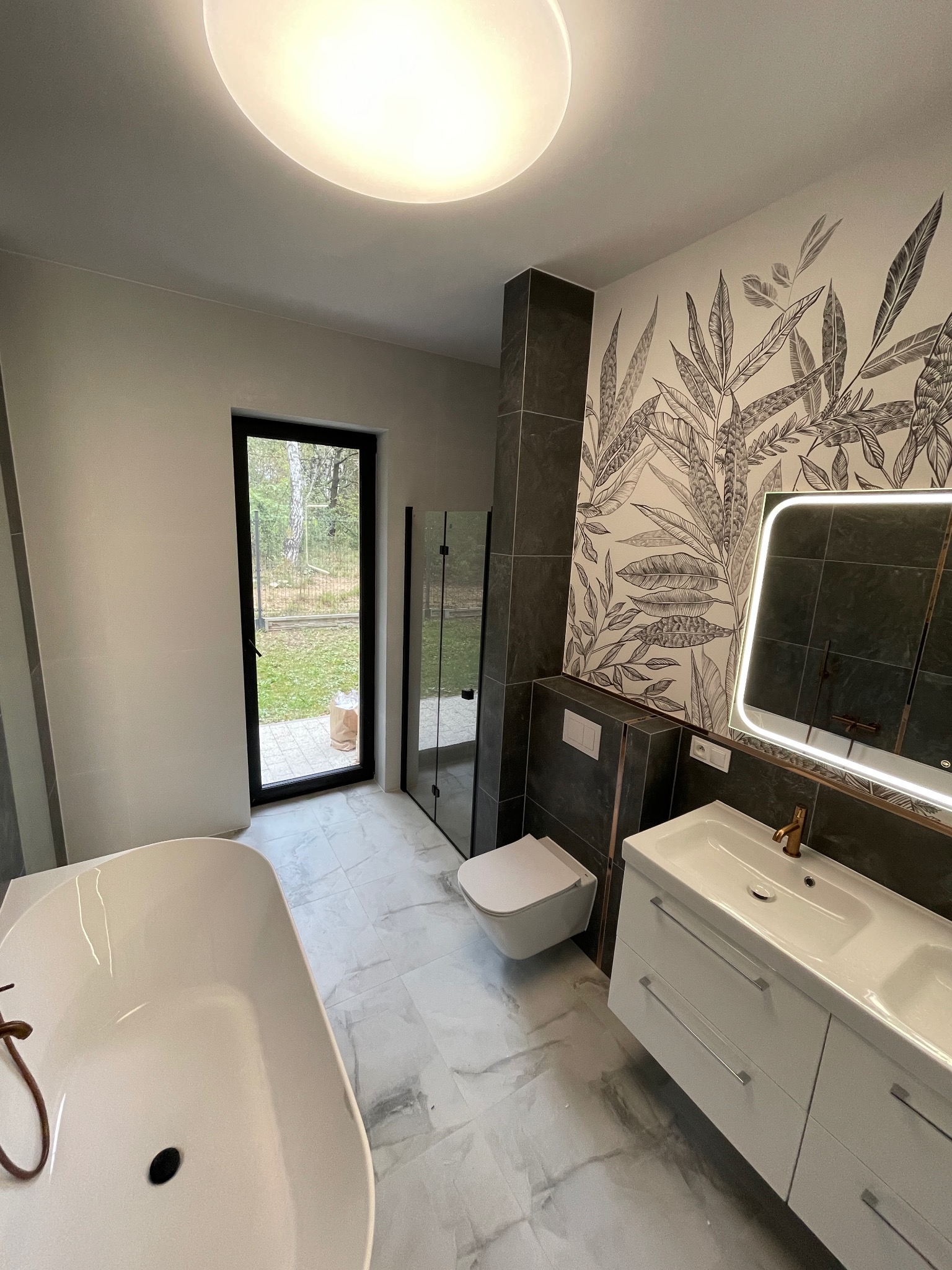 Nowoczesna łazienka z wanną, kabiną prysznicową, umywalką z szafką i toaletą. Na ścianie dekoracyjny motyw roślinny. Widoczne okno z widokiem na zieleń.