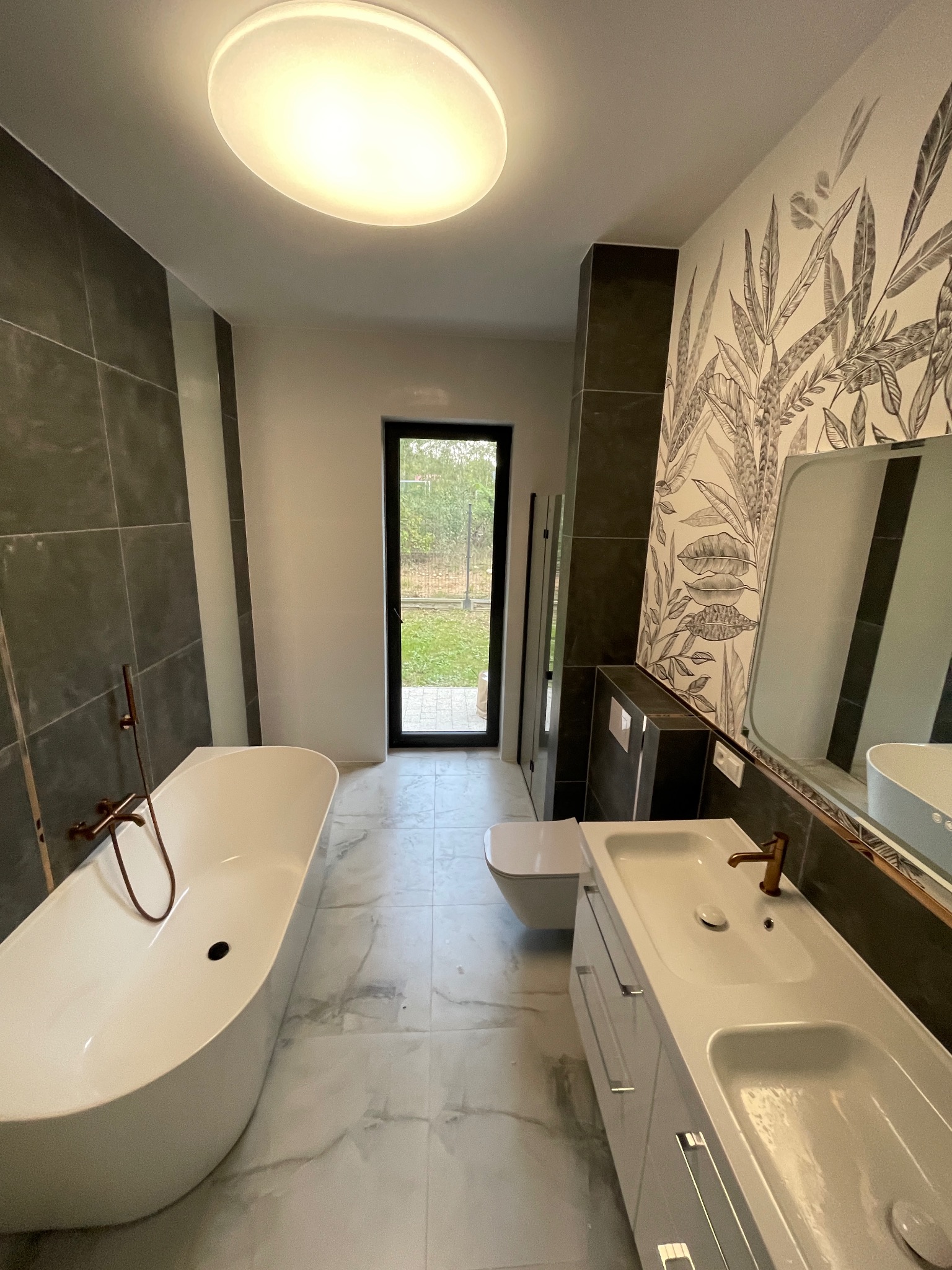 Nowoczesna łazienka z wolnostojącą wanną, umywalką z szafką i toaletą. Ciemne płytki i tapeta z motywem roślinnym. Widok na ogród przez czarne drzwi.