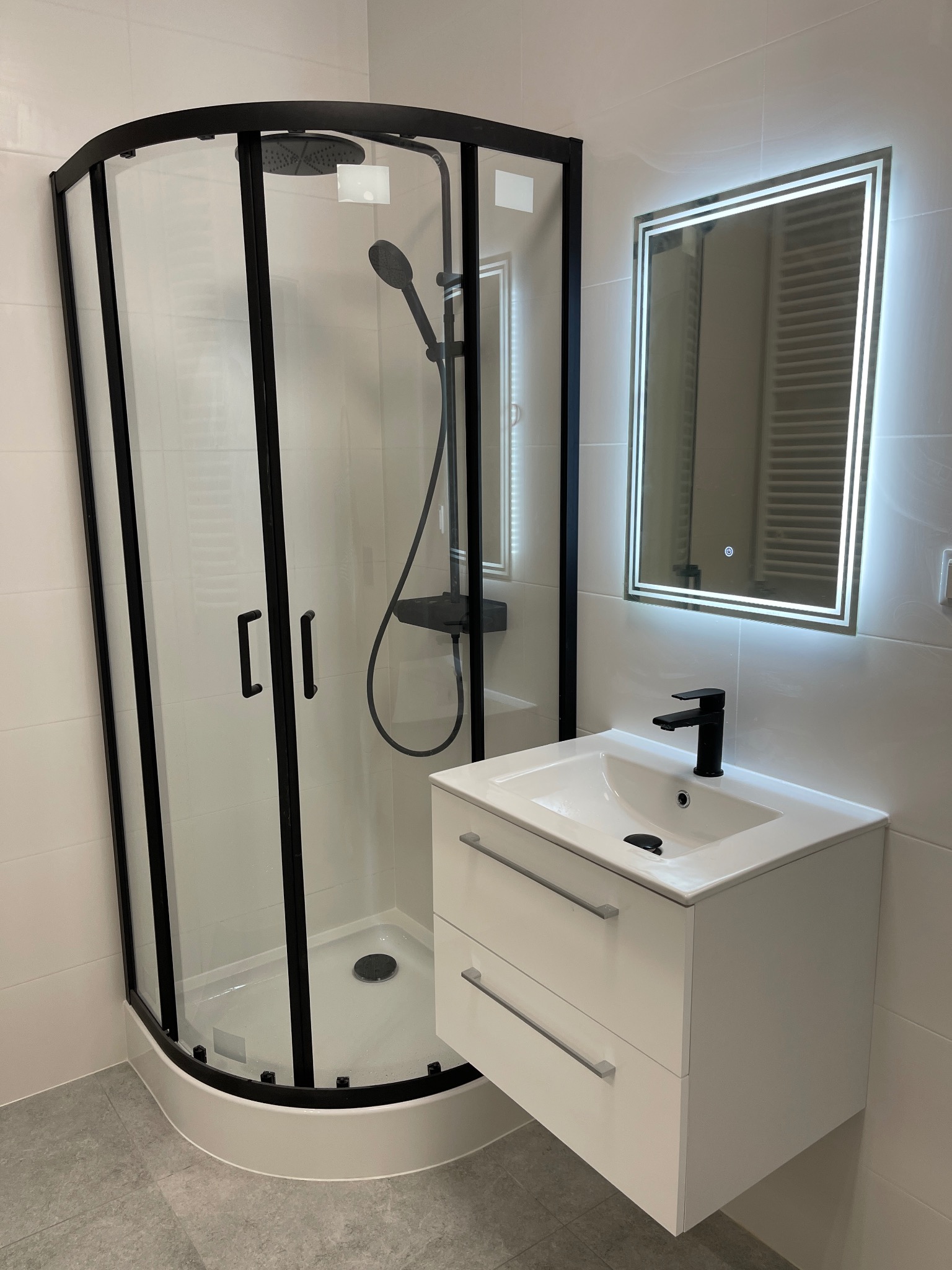 Nowoczesna łazienka z kabiną prysznicową z czarnymi ramami, białą szafką pod umywalką i podświetlanym lustrem. Minimalistyczny design, jasne płytki.