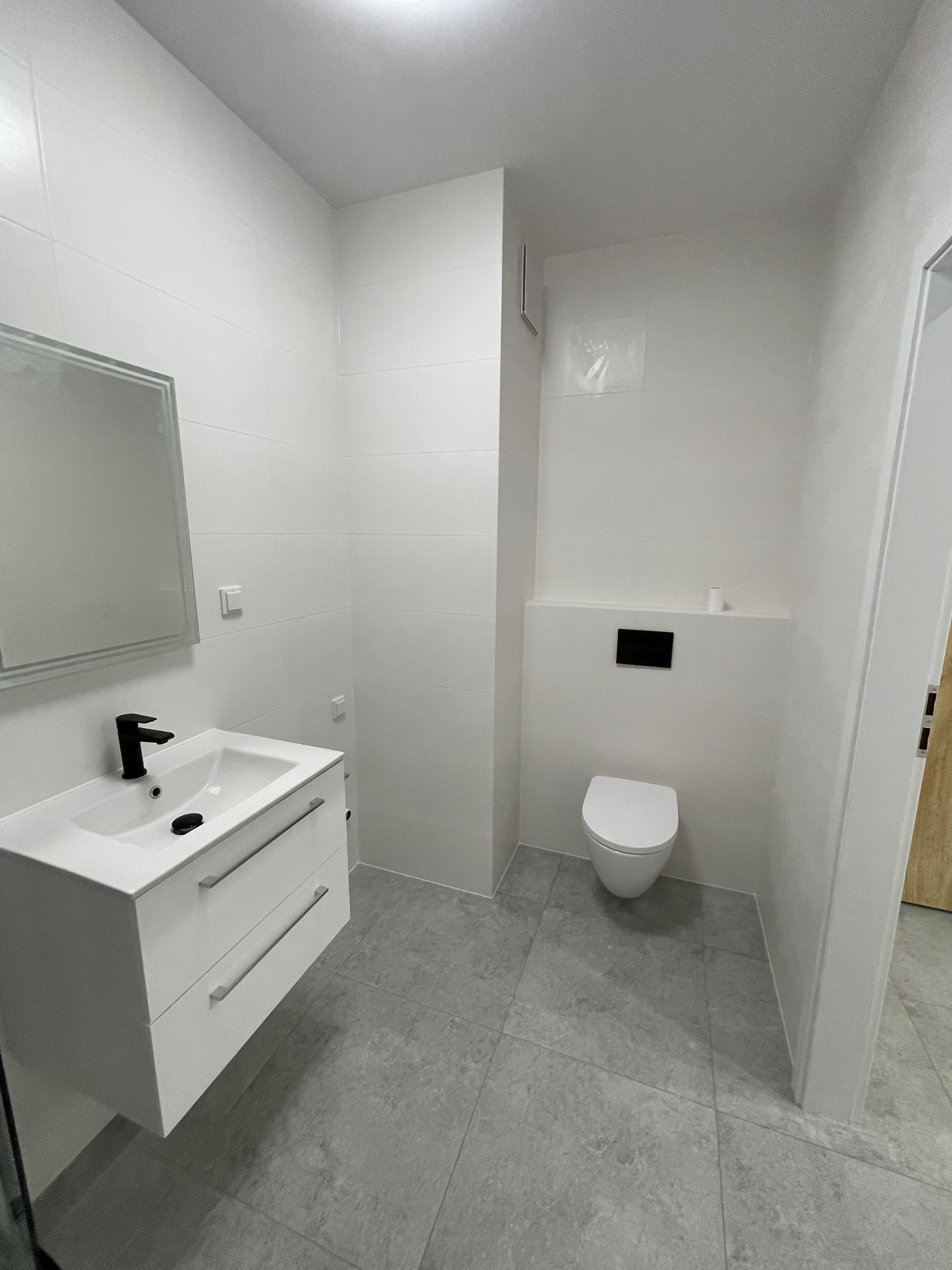 Nowoczesna, minimalistyczna łazienka z białymi płytkami, szarą podłogą i wiszącą toaletą. Umywalka z szafką i czarną armaturą. Całość utrzymana w jasnej tonacji.