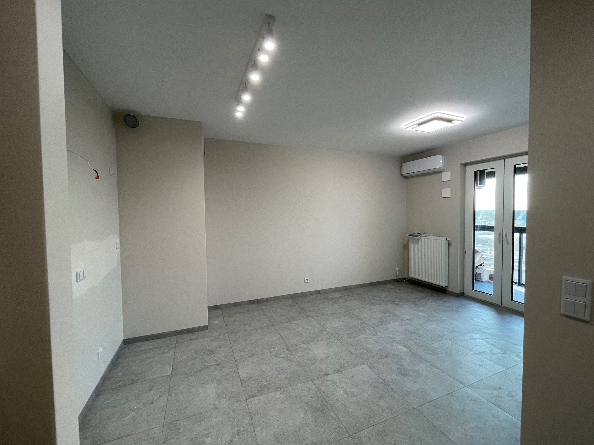 Jasne, przestronne wnętrze z szarą podłogą z płytek, białymi ścianami i oknami balkonowymi. Widoczne oświetlenie sufitowe i grzejnik. Częściowo wykończone ściany.