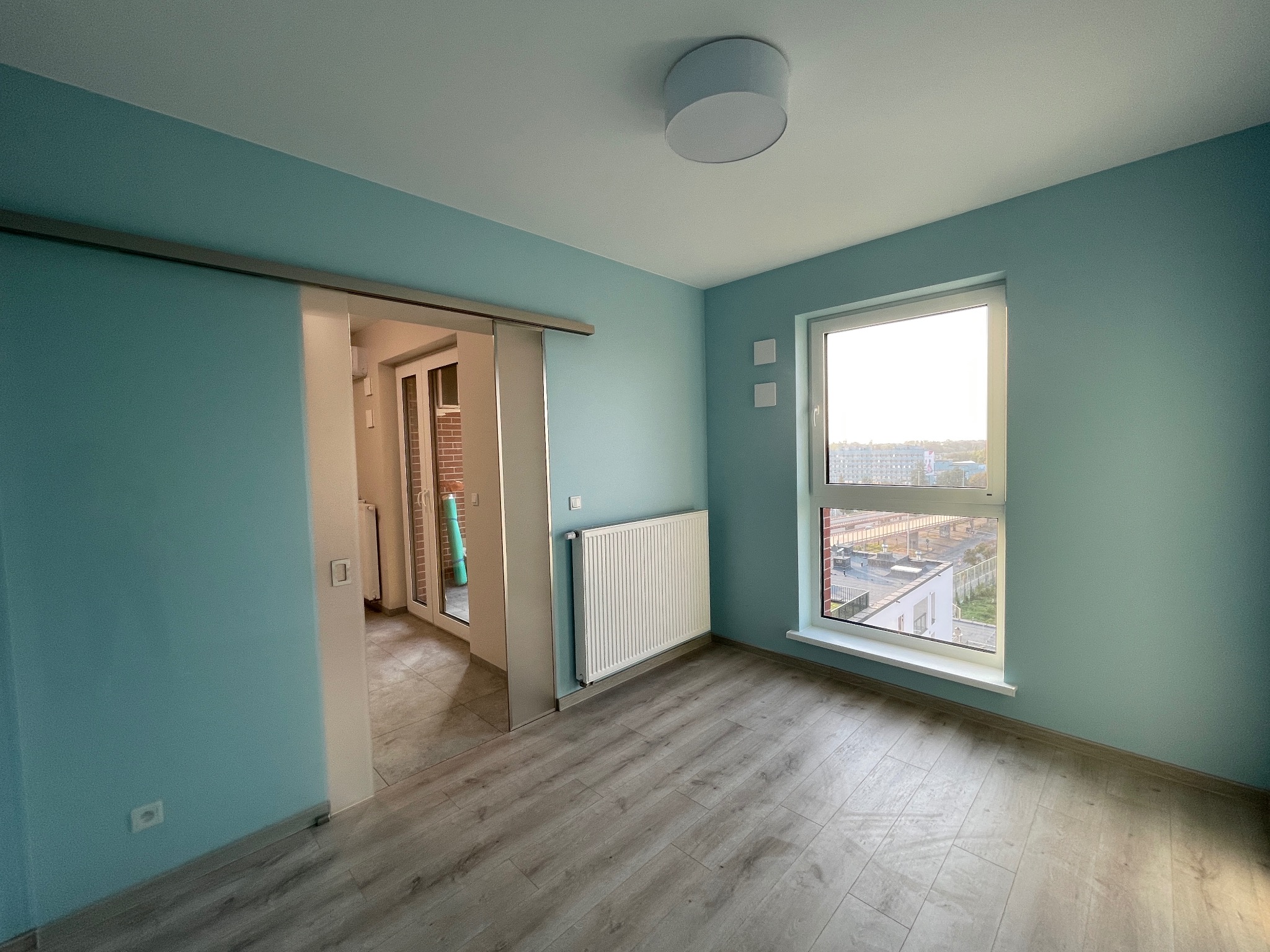 Pomalowane na turkusowo puste wnętrze z panelami, grzejnikiem i oknem z widokiem na miasto. Drzwi przesuwne prowadzą na balkon.
