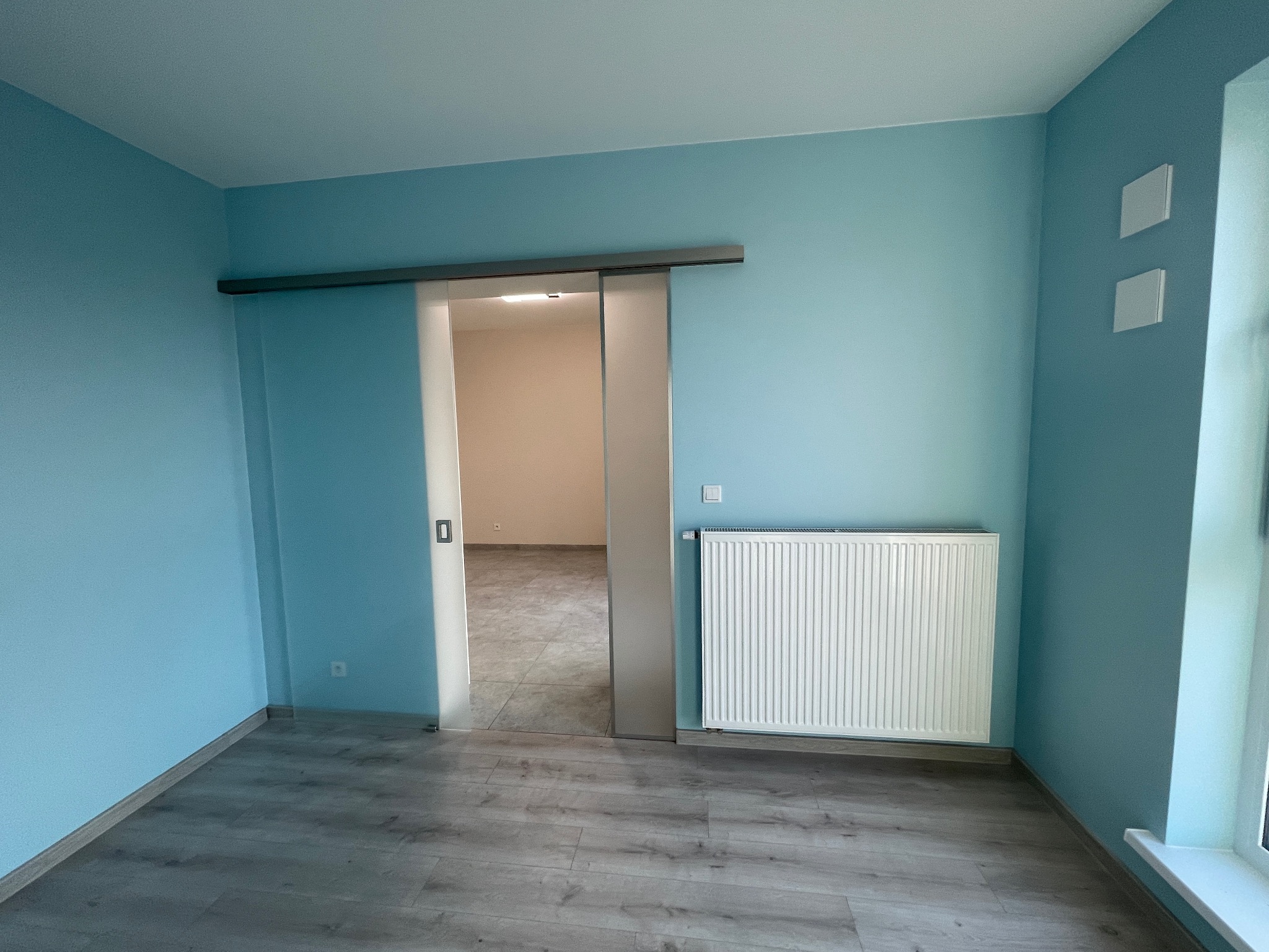 Błękitne wnętrze po remoncie: przesuwne drzwi, grzejnik i panele podłogowe. Widok na sąsiednie pomieszczenie z jasną podłogą.