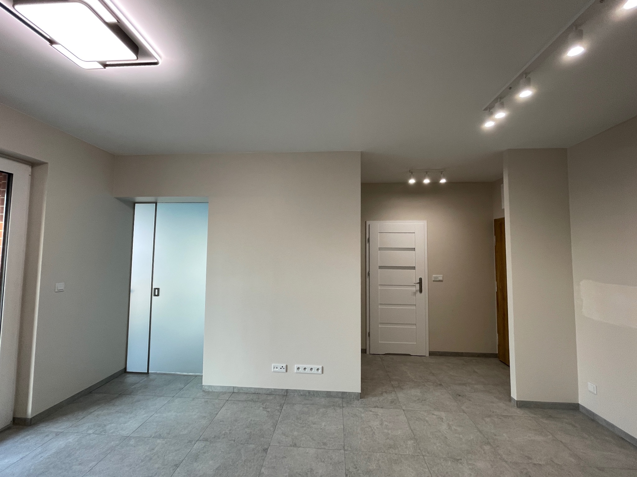 Jasne, przestronne wnętrze po remoncie z szarymi płytkami podłogowymi, przesuwnymi drzwiami i oświetleniem punktowym. Widoczne gniazdka elektryczne.