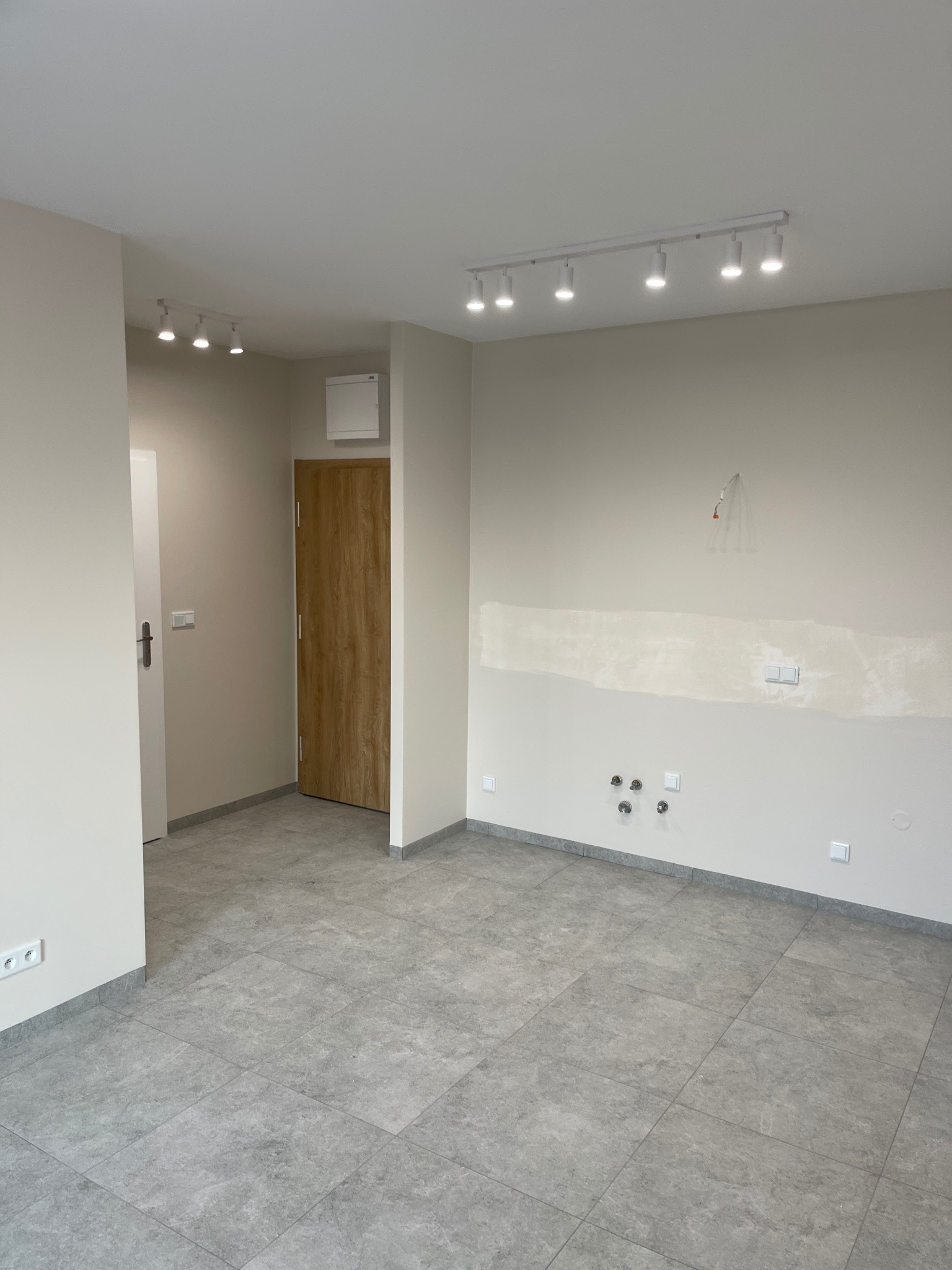 Jasne, wykończone wnętrze z szarą podłogą z płytek, drewnianymi drzwiami i białym oświetleniem sufitowym. Ściany w odcieniach beżu, widoczne gniazdka i instalacje.