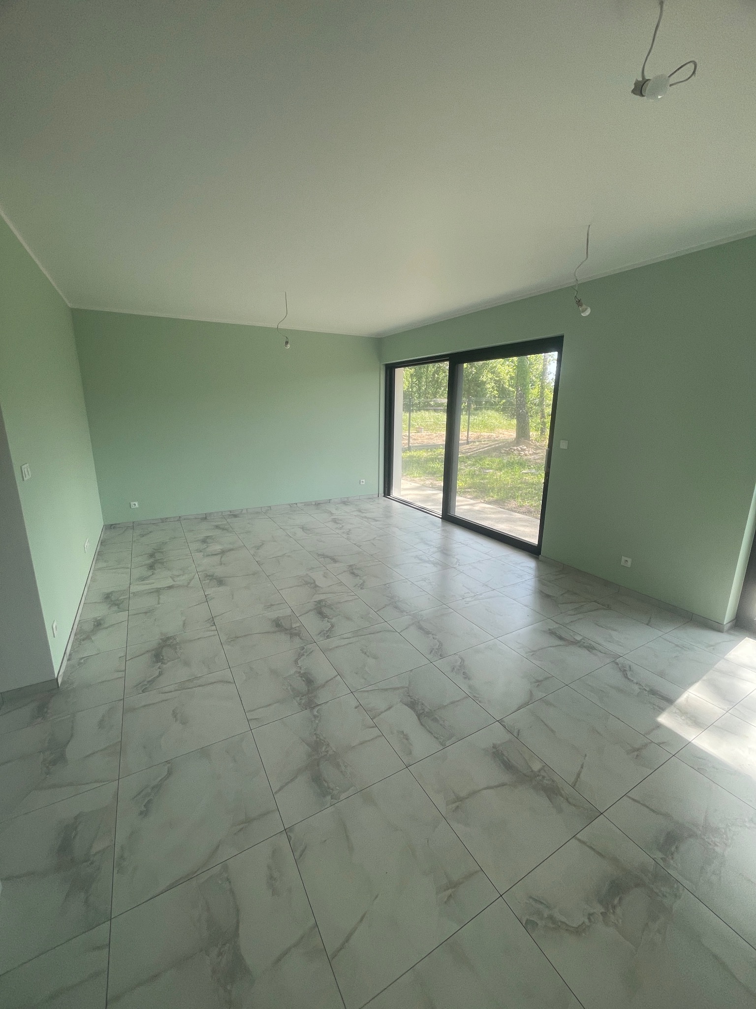 Jasne wnętrze z zielonymi ścianami i marmurową podłogą. Duże okno z widokiem na zieleń. Minimalistyczny styl, surowe żarówki. Świeżo po remoncie, gotowe do umeblowania.