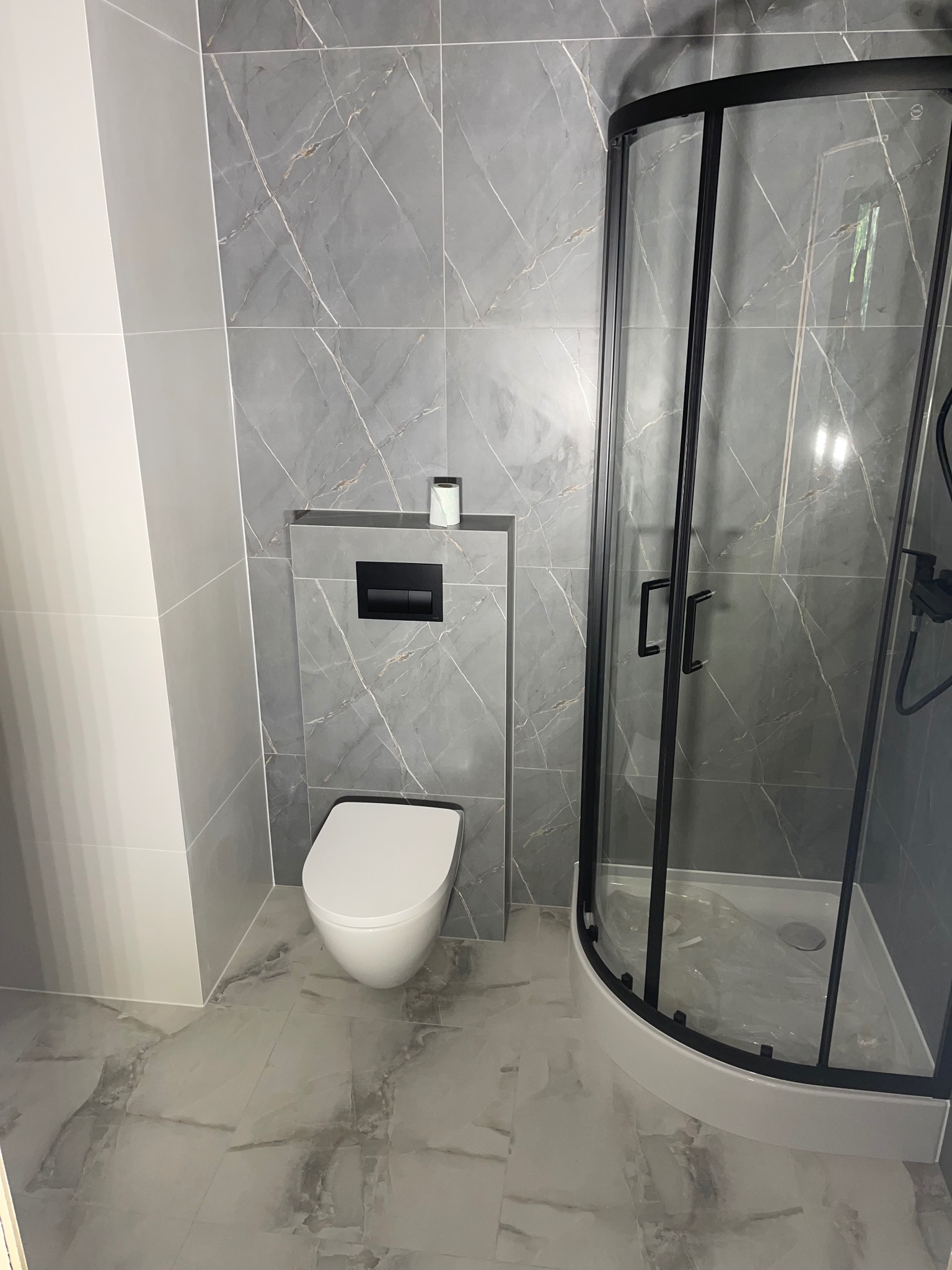 Nowoczesna łazienka z szarymi płytkami imitującymi marmur, kabiną prysznicową z czarnymi ramami i podwieszaną toaletą. Minimalistyczny design, widok na całe pomieszczenie.