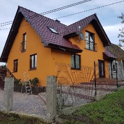 Łukasz Zawadziński - Dwupiętrowy dom z pomarańczową elewacją i ciemnoczerwonym dachem, widoczny zza ogrodzenia. Na ścianie drabina, a na dachu okno. Widoczne elementy wykończenia.